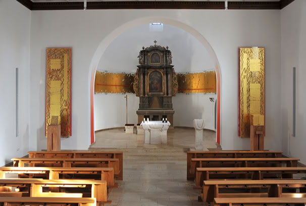 Katholische Pfarrkirche St. Josef, Herzogenaurach-Niederndorf. Chorraumgestaltung. Glasband, 10 Segmente aus 8mm Floatglas, Optiwhite, mit Abstand vor die Wand montiert, vergoldet mit 24 Karat Blattgold.  Bildtafeln (jeweils mit Tabernakel und Evangeliar): je zwei mit Abstand hintereinander geschichtete Floatglasscheiben, Optiwhite, rückseitiger Farbdruck, vorderseitig Blattvergoldung.  Stelen für Tabernakel und Evangeliar: Roteiche gesägt (Till Hausmann).  Tabernakel: Floatglas, Optiwhite, außenseitig rot lackiert unter Blattvergoldung 24 Karat.